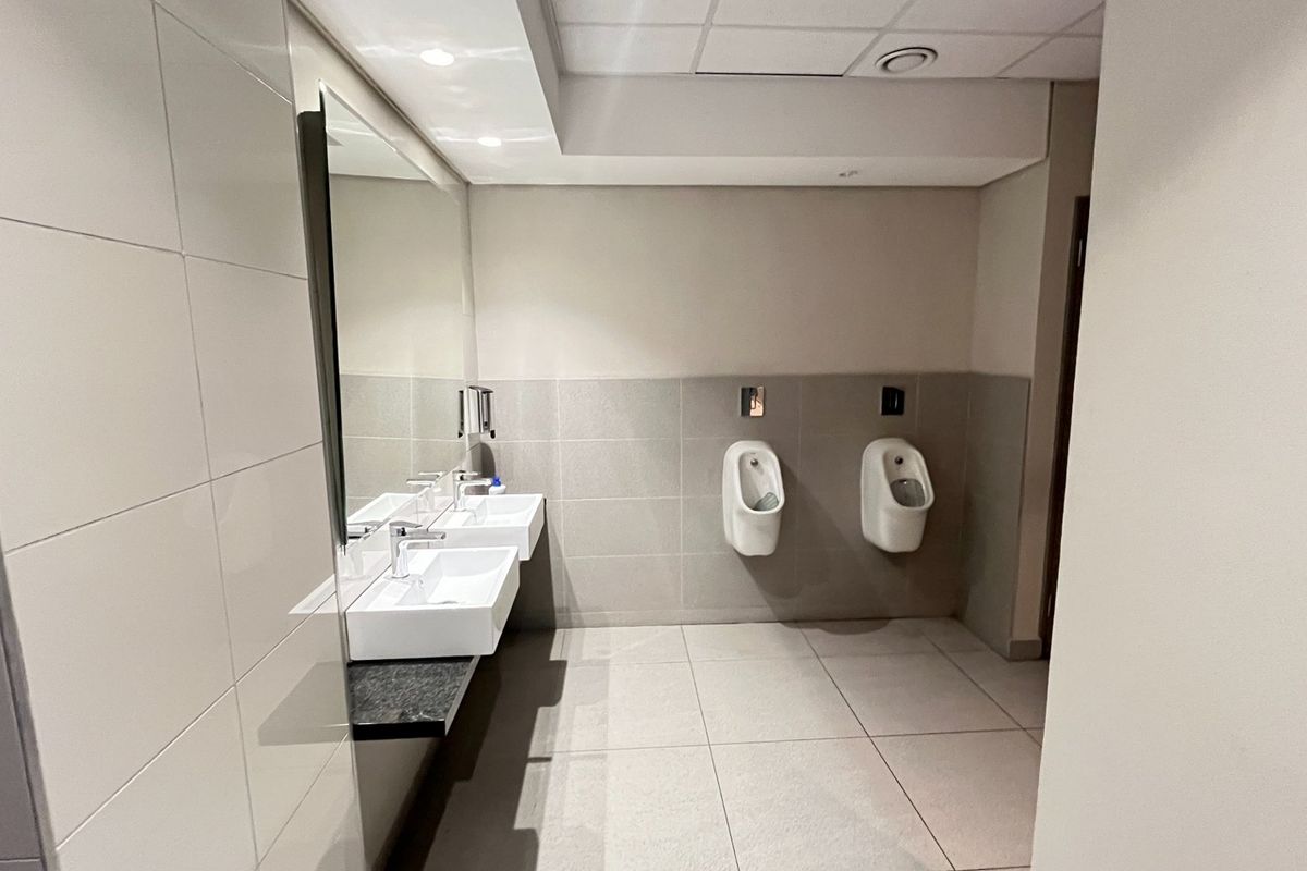A clean, modern commercial restroom featuring wall-mounted urinals, dual ceramic washbasins, large wall mirror, and neutral tiled finishes.