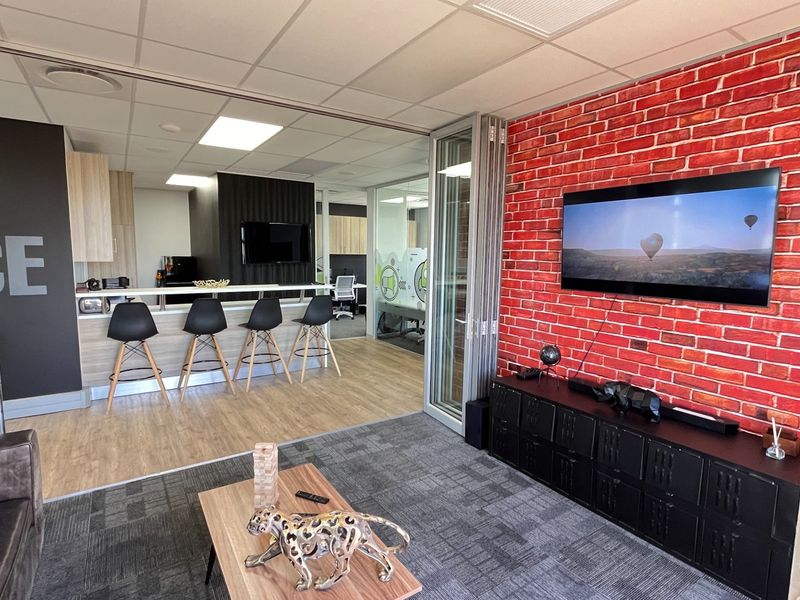 Modern open-plan commercial office space featuring a striking red brick accent wall, kitchenette area with breakfast bar, and flexible bi-fold glass partitions.