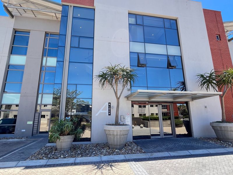Modern commercial office building entrance featuring a glass curtain wall facade, metal framing, and a paved approach with integrated landscaping.