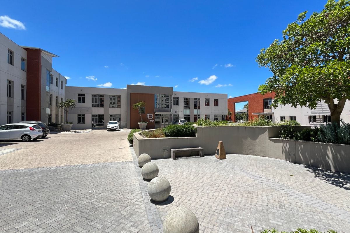 A well-maintained commercial courtyard featuring paved surfaces, concrete bollards, landscaped garden beds, and access to a multi-story office building complex.