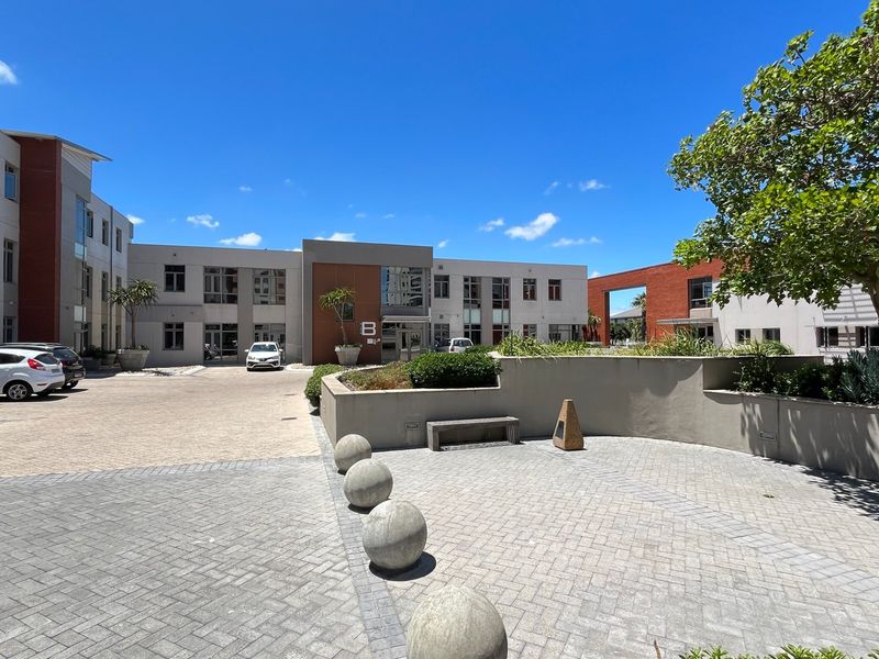 A well-maintained commercial courtyard featuring paved surfaces, concrete bollards, landscaped garden beds, and access to a multi-story office building complex.