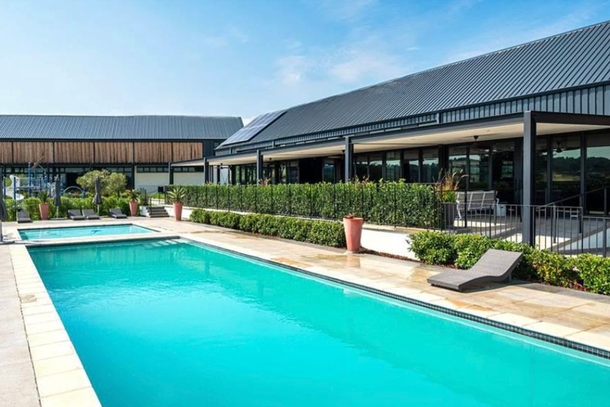 A well-maintained swimming pool area featuring a paved deck, landscaped hedging, and a modern building with a metal roof and covered patio.