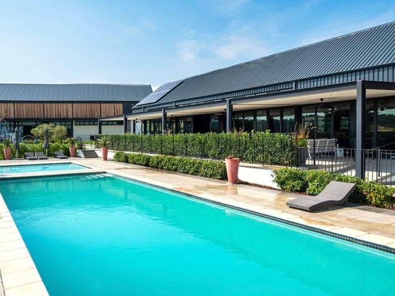 A well-maintained swimming pool area featuring a paved deck, landscaped hedging, and a modern building with a metal roof and covered patio.