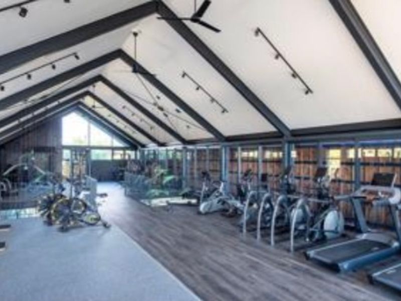 A spacious, modern gym featuring a high vaulted ceiling with exposed steel beams, track lighting, and extensive glass windows providing natural light.