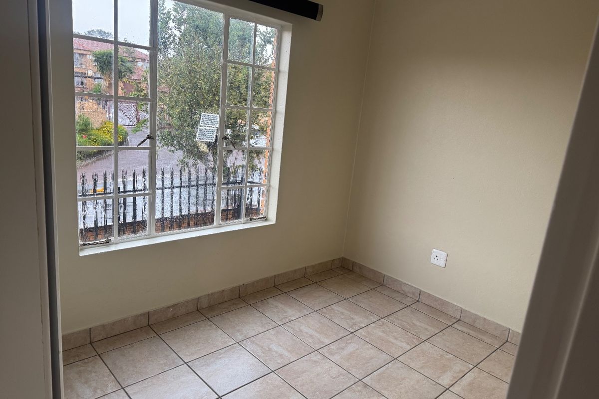 A bright, empty bedroom featuring neutral tiled flooring and a large window providing natural light and a view of the complex exterior.