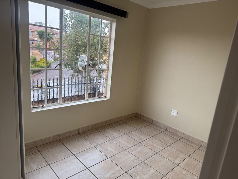 A bright, empty bedroom featuring neutral tiled flooring and a large window providing natural light and a view of the complex exterior.