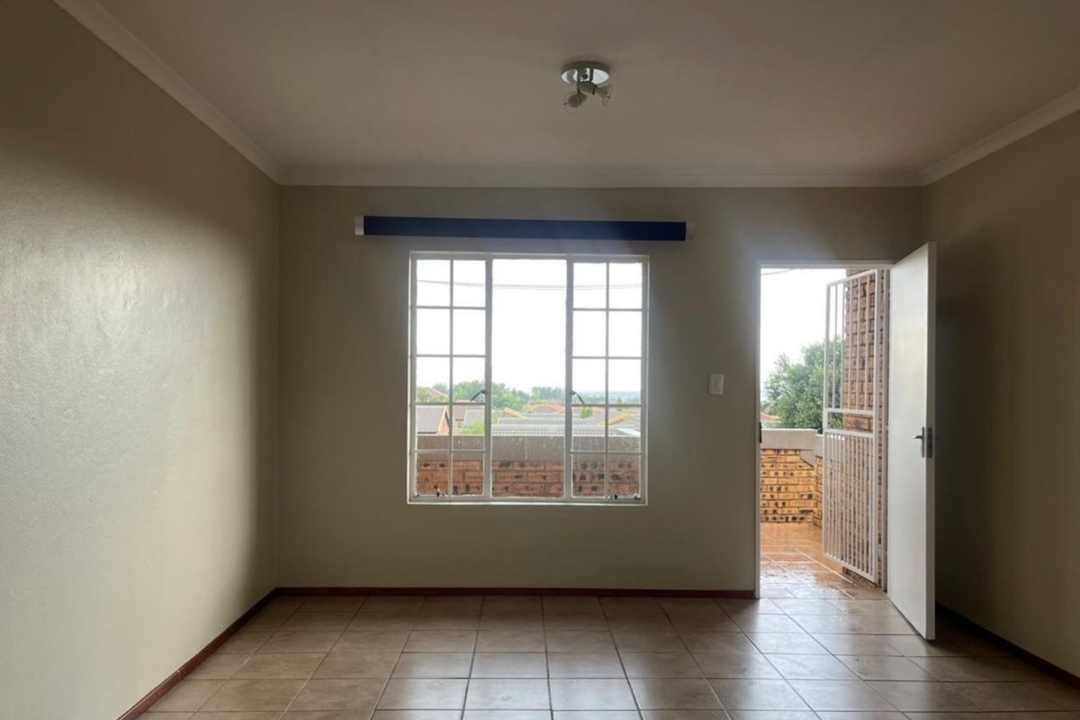 An empty, neutrally painted living room featuring tiled flooring, a large window with security bars, and direct access to an outdoor balcony area.