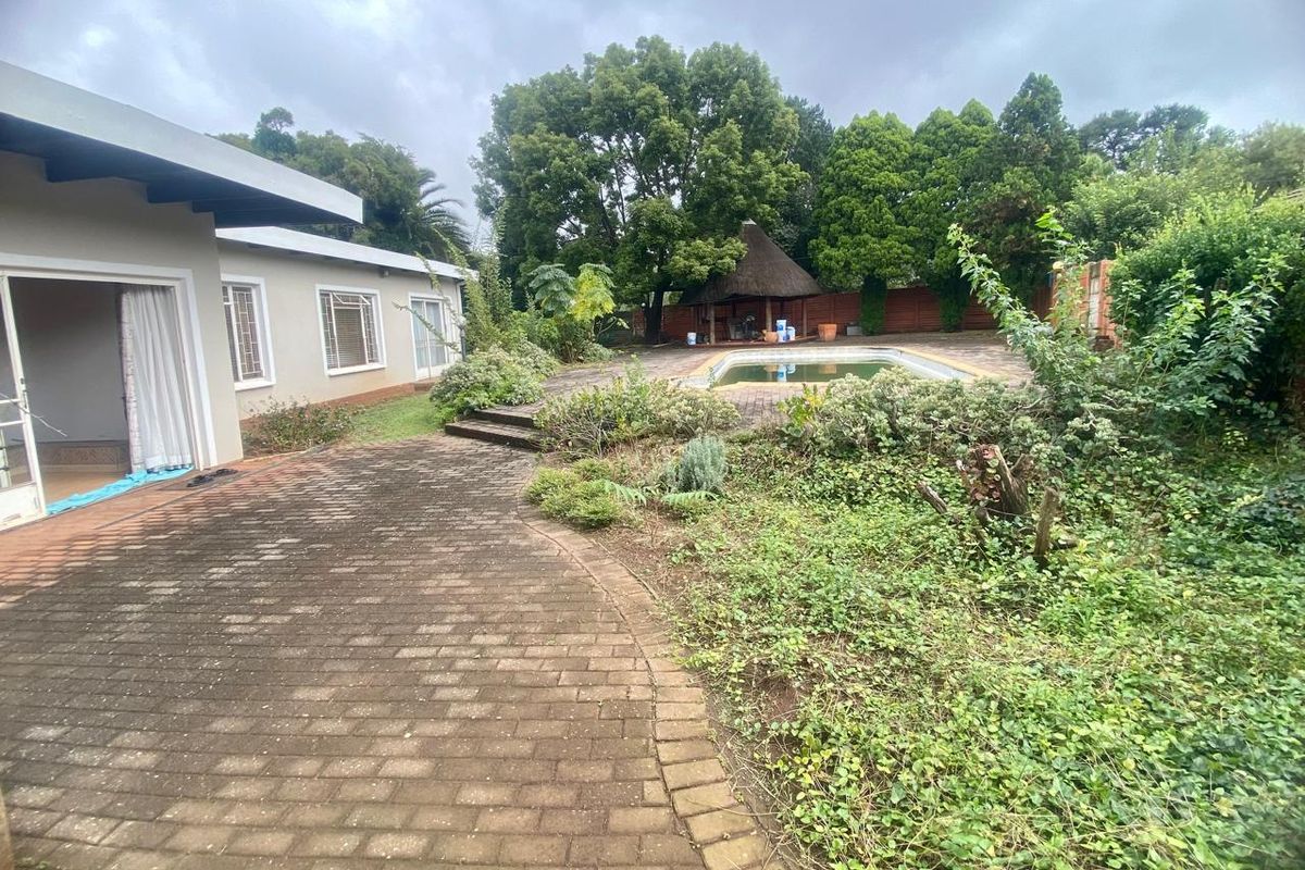 Rear exterior view featuring a swimming pool, a thatched lapa, and a paved patio area surrounded by an overgrown garden.