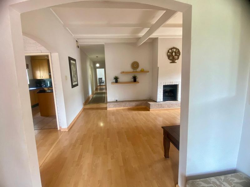 Spacious living room featuring laminate flooring, exposed ceiling beams, and a fireplace with a tiled hearth, leading into a kitchen and hallway.