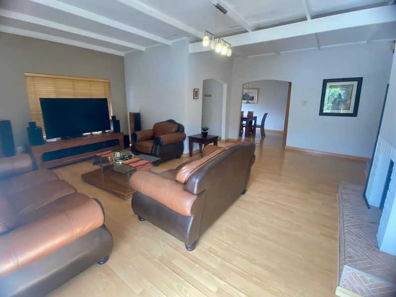 Spacious living room featuring exposed beam ceilings, light laminate flooring, and a brick fireplace hearth, with an open archway leading to the dining area.