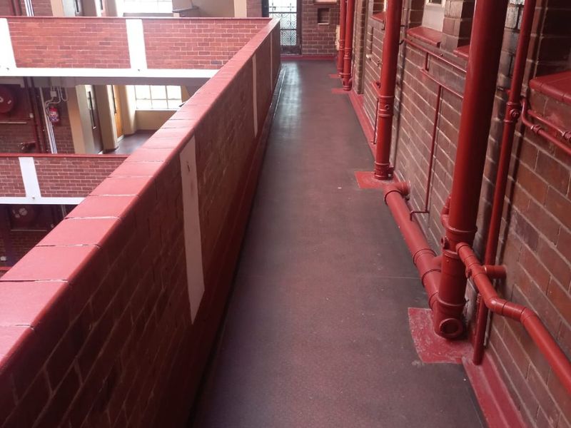 An open-air apartment building walkway featuring exposed brick walls, red painted piping, and a brick balustrade overlooking the central courtyard.