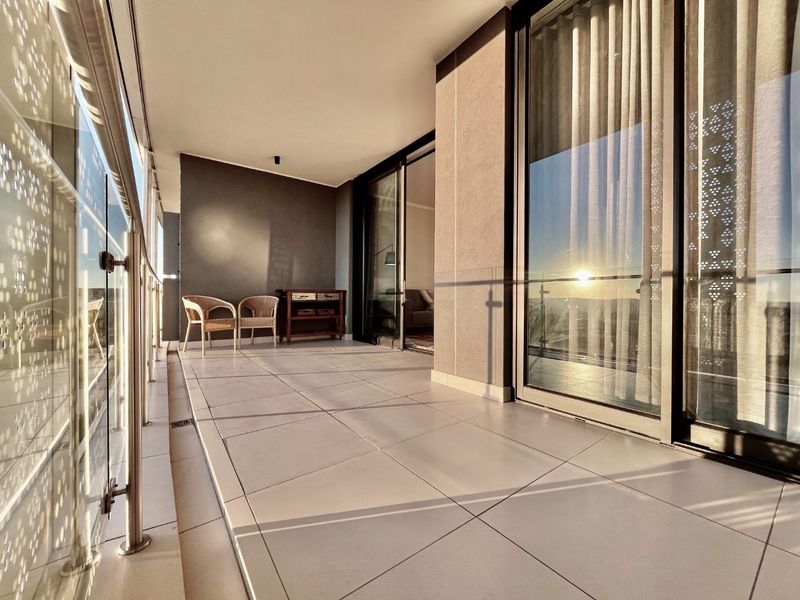 A spacious, modern balcony featuring large format floor tiles, a sleek glass balustrade, and expansive sliding glass doors providing seamless indoor-outdoor flow.