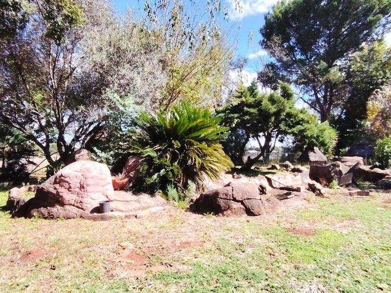 A landscaped garden area featuring large natural rock formations, mature trees, and established shrubbery on a farm property.