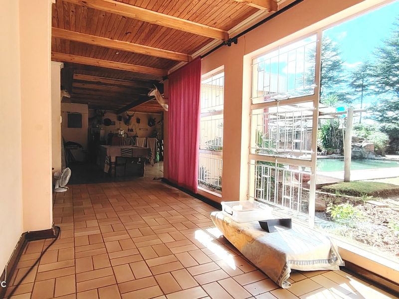 A sunlit living area featuring large windows with views of the garden and pool, finished with tiled flooring and a rustic timber ceiling.