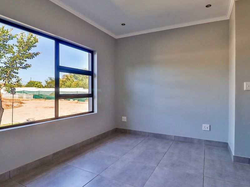 A bright, empty bedroom featuring modern grey floor tiles, neutral walls, and a large aluminium window providing natural light.