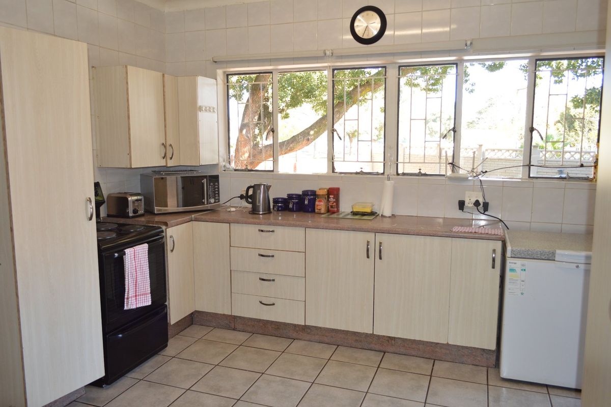 A functional kitchen featuring light-toned built-in cabinetry, tiled splashbacks, and tiled flooring, with large windows providing natural light and security bars.