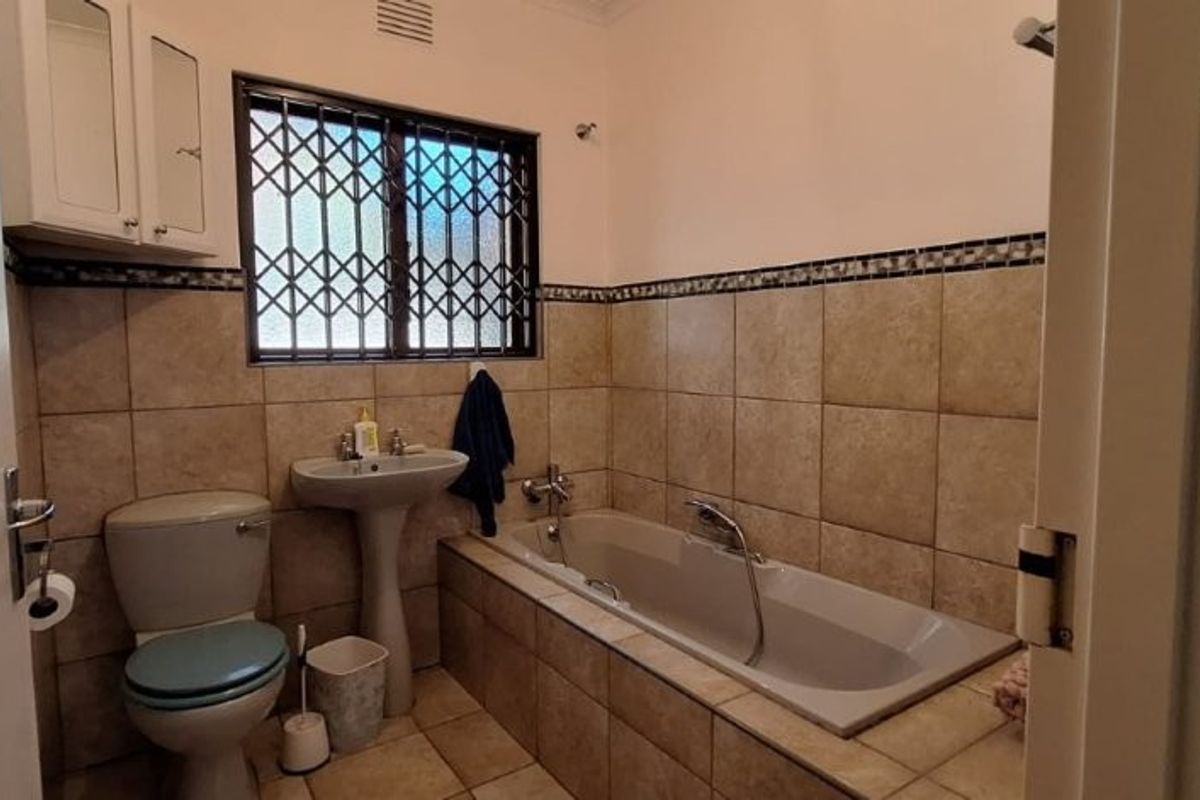 A functional bathroom featuring a built-in bathtub, pedestal basin, toilet, and tiled finishes, complete with security bars on the window.