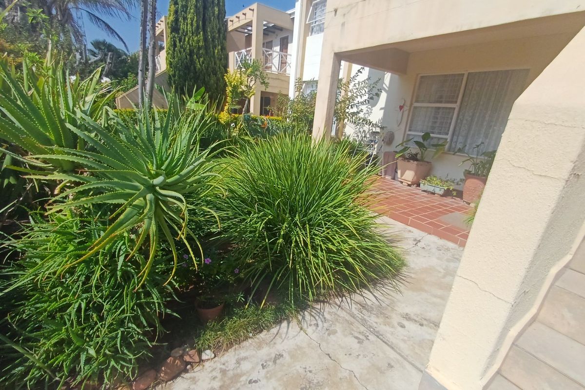 A rear exterior view of the apartment featuring a tiled patio area, concrete walkway, and established garden landscaping.