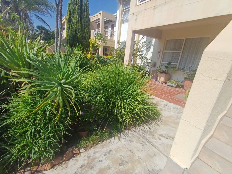 A rear exterior view of the apartment featuring a tiled patio area, concrete walkway, and established garden landscaping.