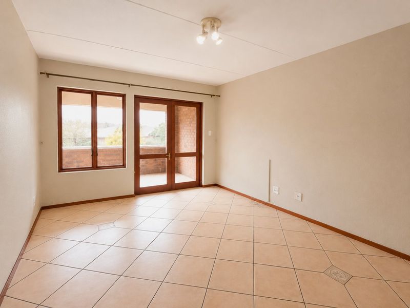 A bright, empty living room featuring neutral tiled flooring, a ceiling light fixture, and direct access to an outdoor balcony through glass-paneled doors.