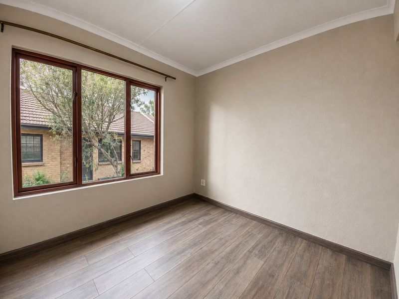 A bright, empty bedroom featuring modern wood-look laminate flooring, neutral wall finishes, and a large window with a view of the exterior brick building.