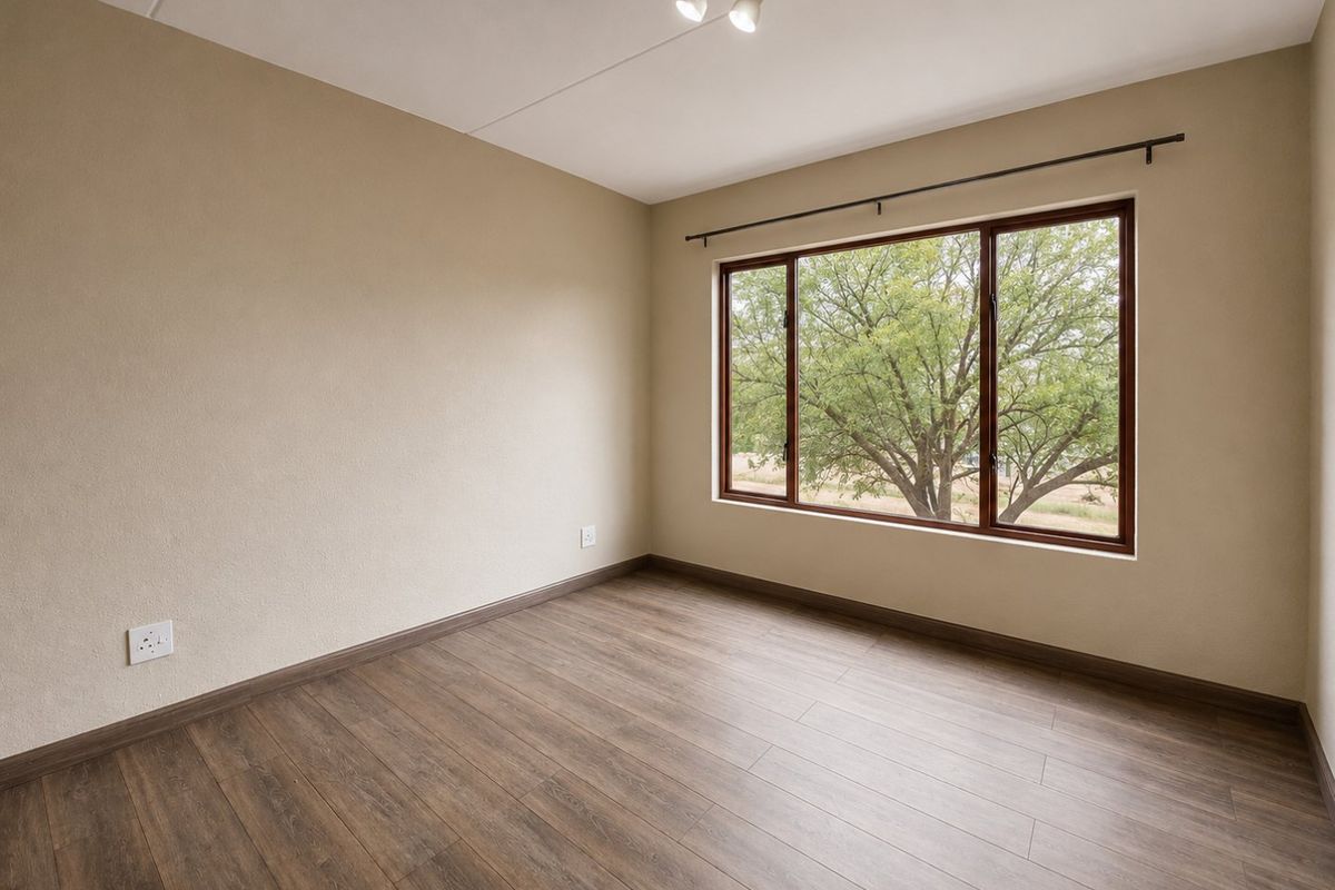 A bright, empty bedroom featuring wood-look laminate flooring, neutral walls, and a large window with a view of mature trees.