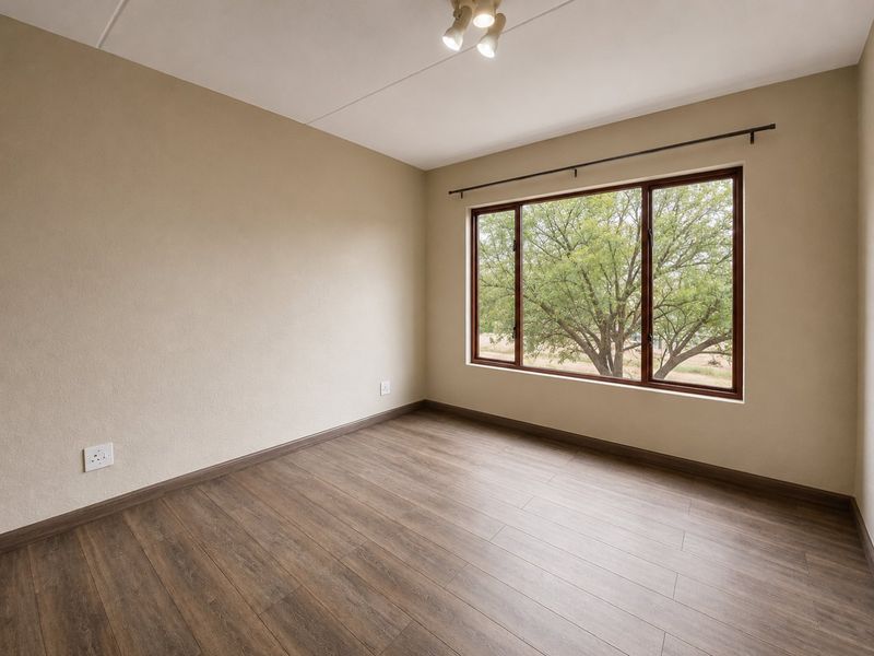 A bright, empty bedroom featuring wood-look laminate flooring, neutral walls, and a large window with a view of mature trees.
