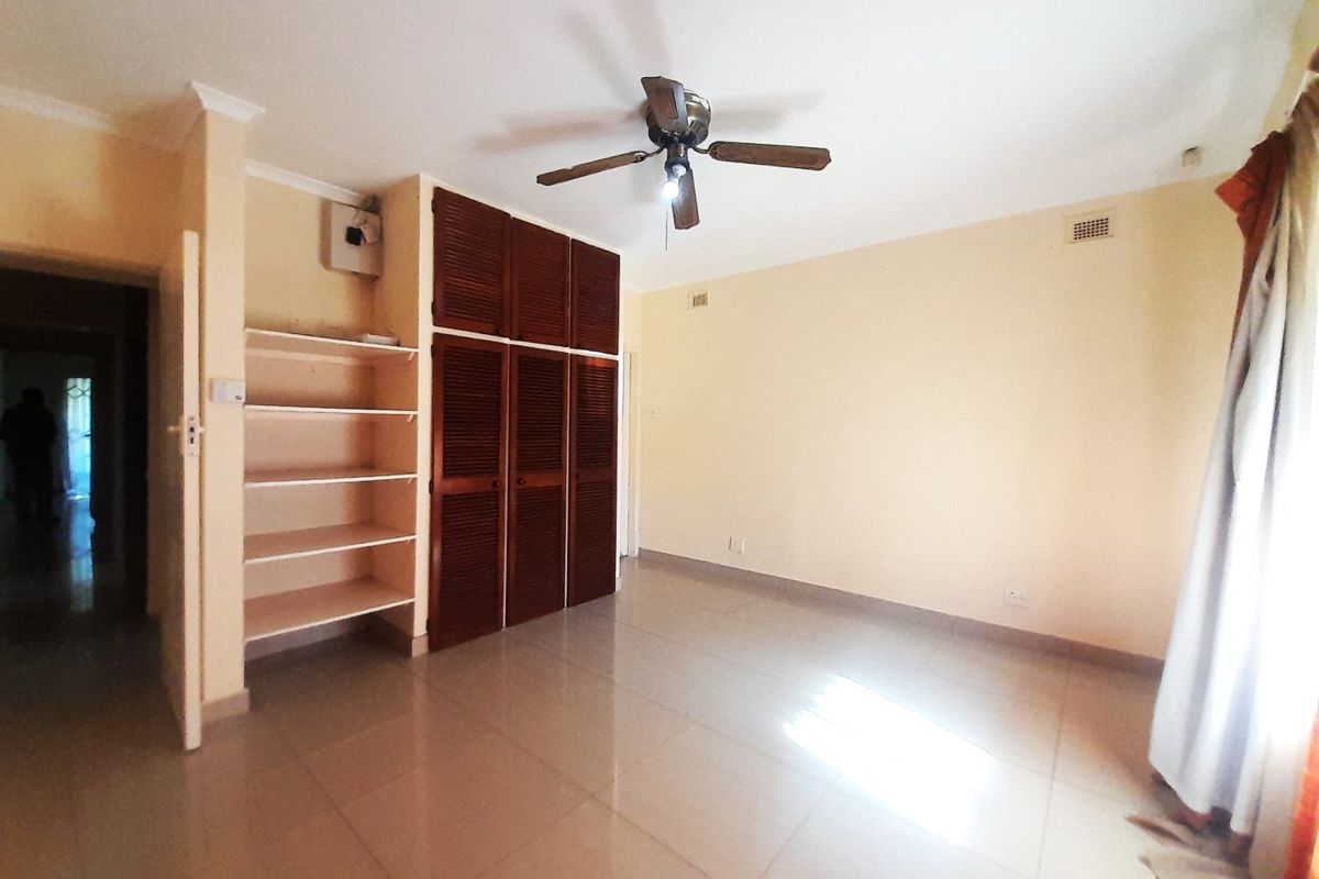 A spacious bedroom featuring tiled flooring, built-in wooden cupboards with integrated shelving, and a ceiling fan.