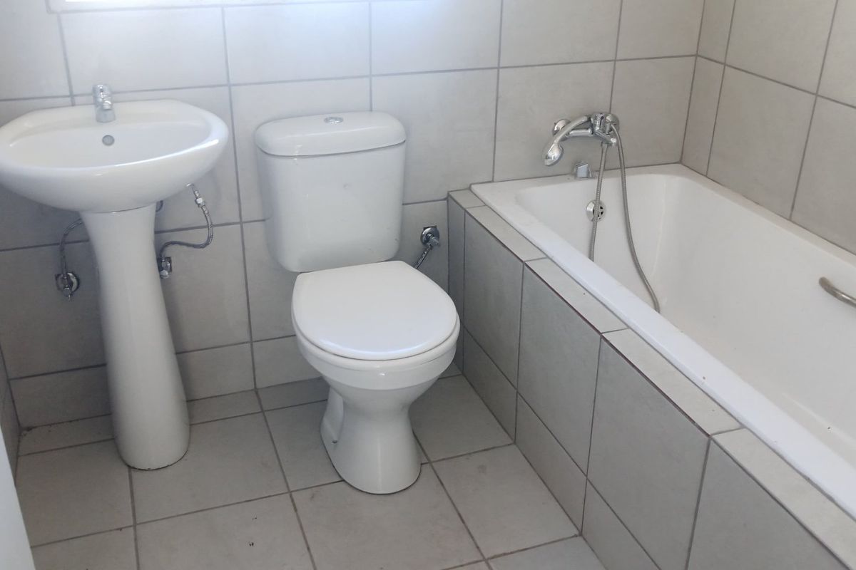 A functional bathroom featuring a pedestal washbasin, standard toilet, and a bathtub with a shower mixer, finished with neutral wall and floor tiles.