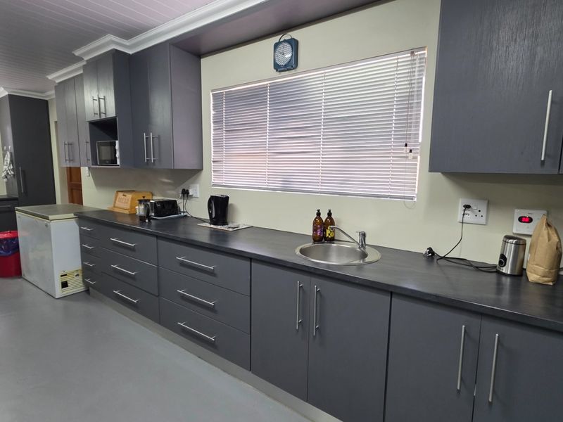 A functional kitchen featuring dark grey cabinetry, laminate countertops, a stainless steel sink, and neutral wall finishes with venetian blinds.