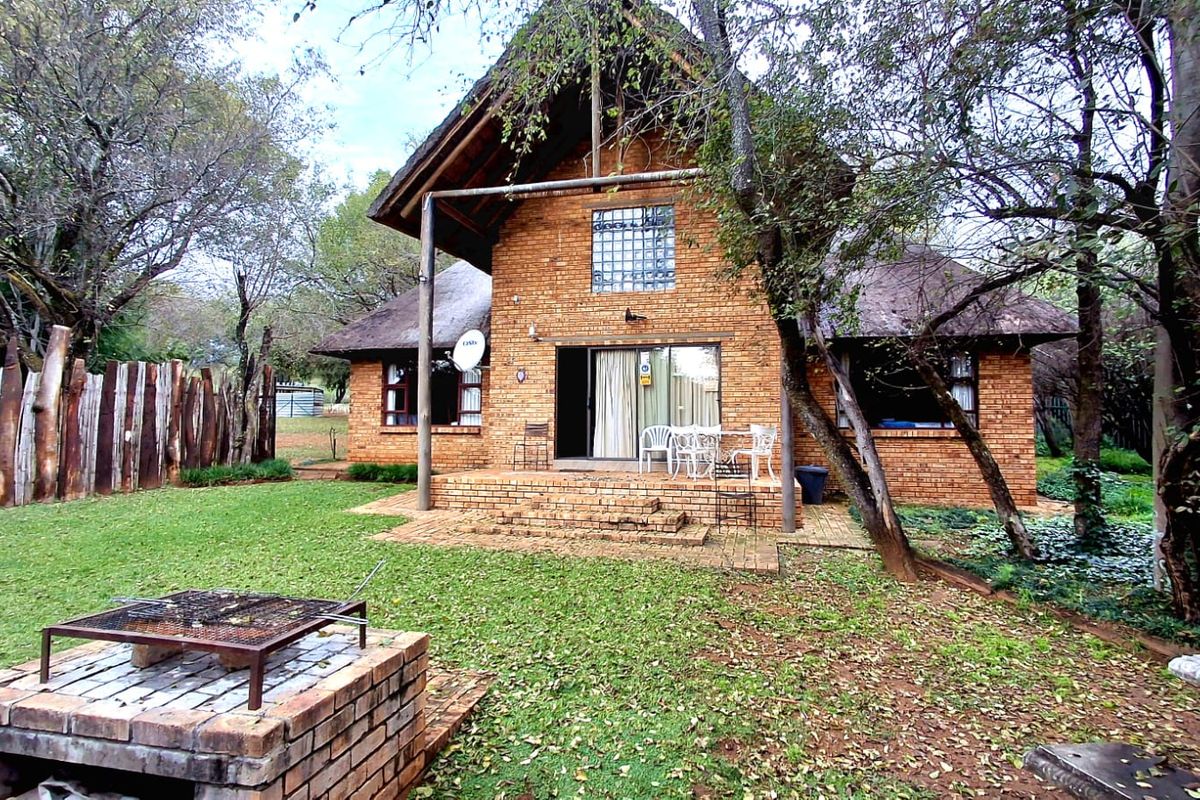Rear view of a rustic brick farmhouse featuring a thatched roof, a paved patio area, and a built-in brick braai set within a garden.