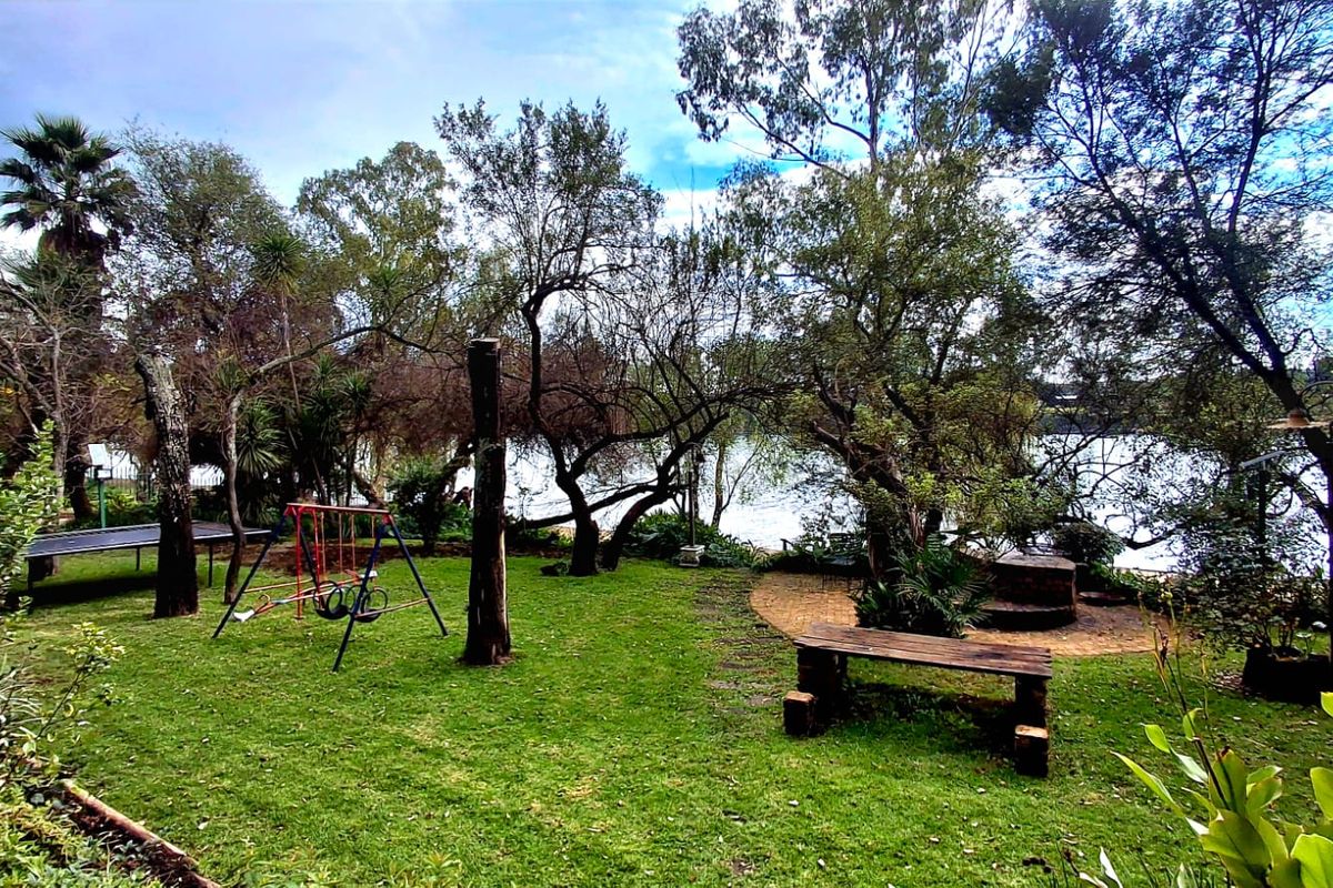 A lush, mature garden featuring established trees, a manicured lawn, and a paved seating area overlooking the water.