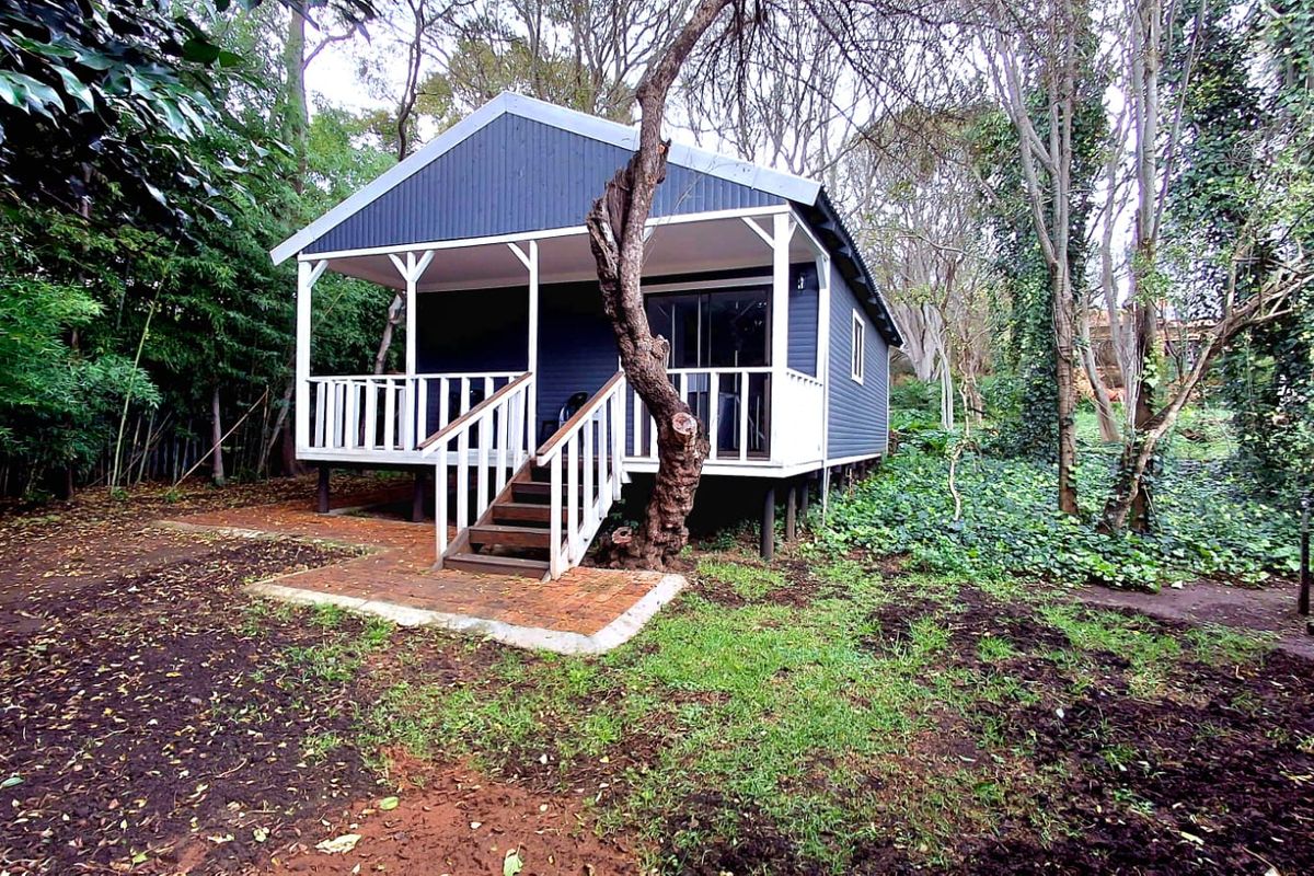 A detached cabin featuring dark corrugated metal cladding, a raised front deck with white balustrades, and a brick-paved entrance set within a wooded garden.