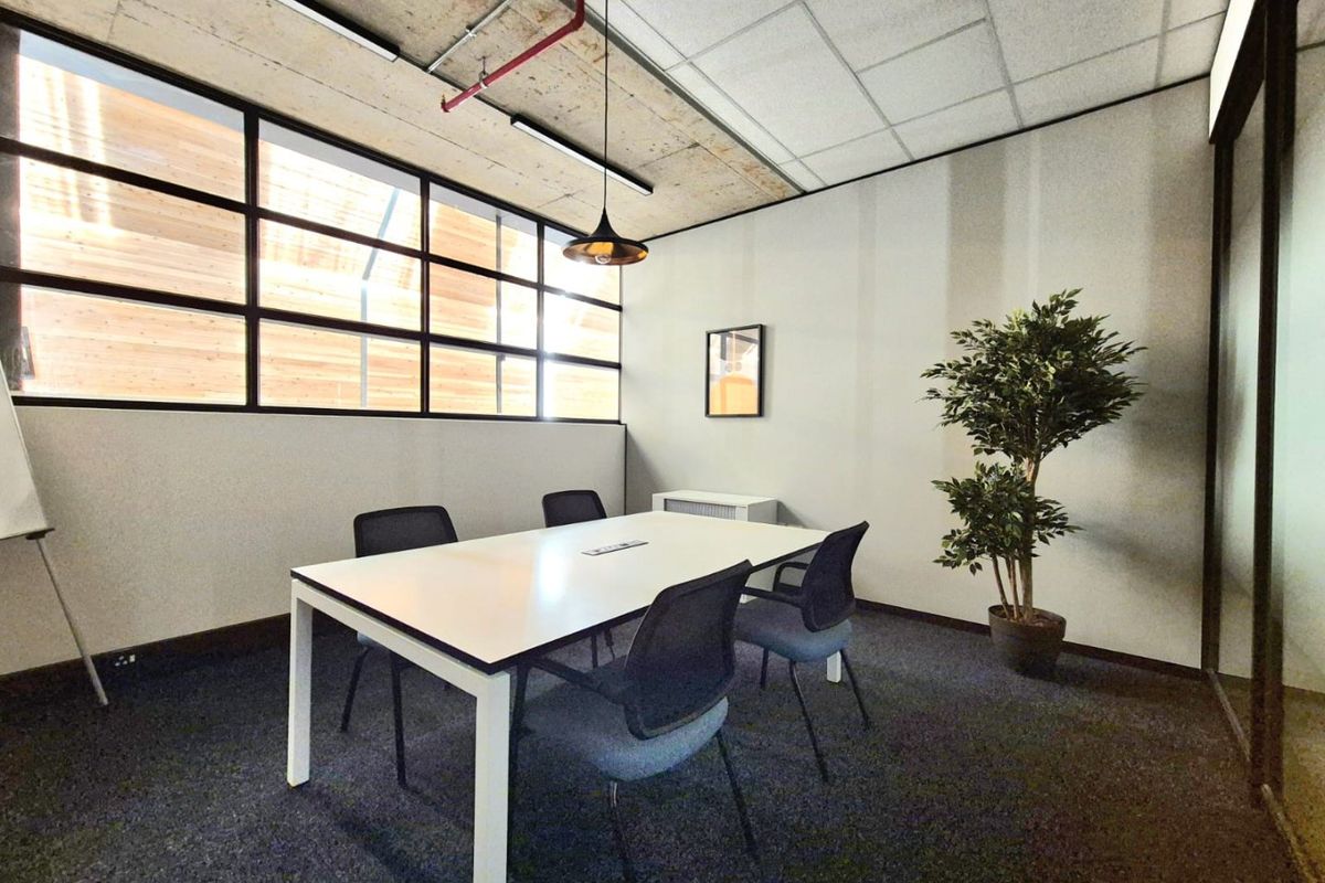 A modern office meeting room featuring an exposed concrete ceiling, industrial pendant lighting, and large grid-style windows providing natural light.