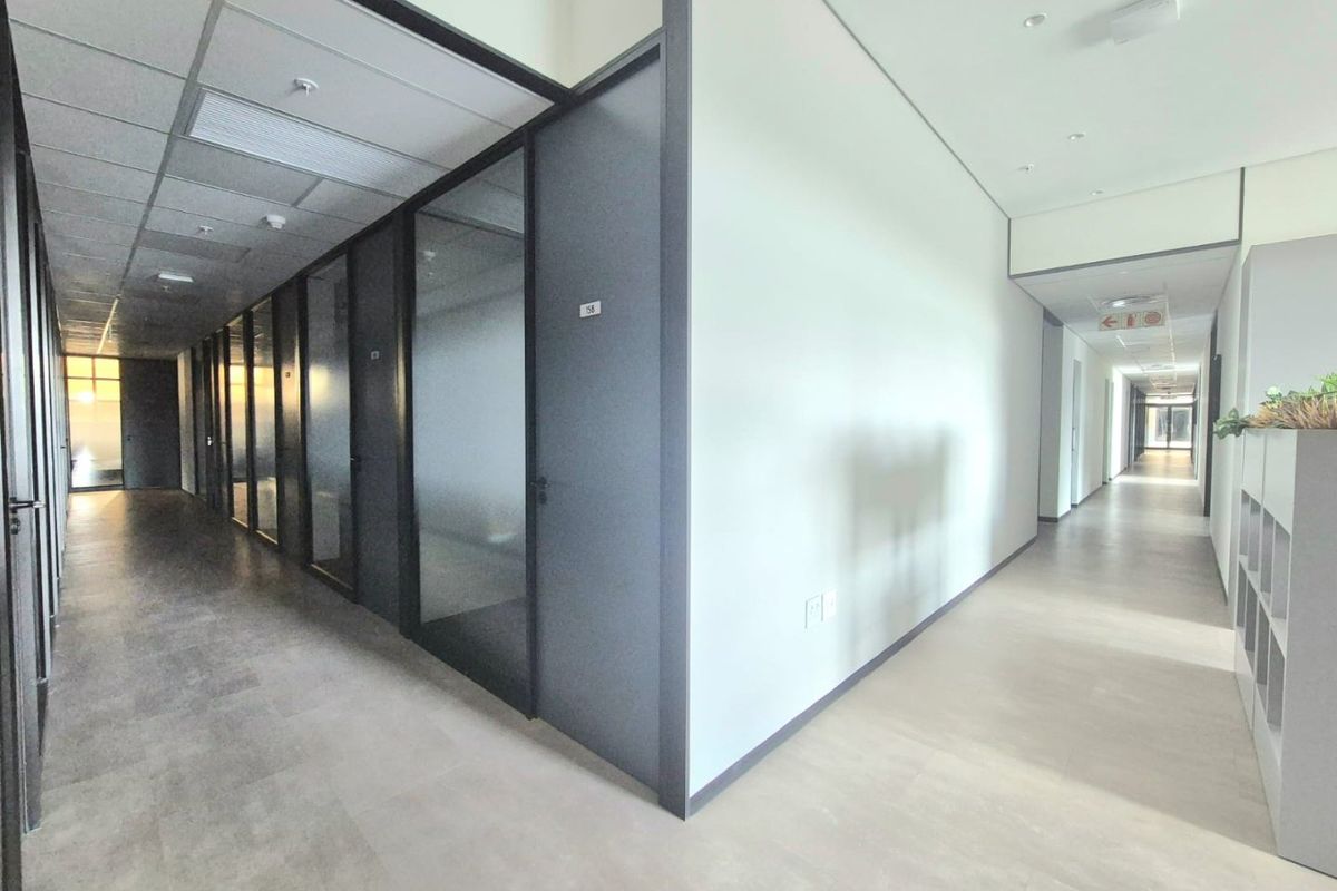 A modern commercial office hallway featuring glass-partitioned offices, neutral vinyl flooring, and a clean, professional aesthetic with recessed lighting.