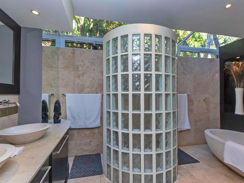 Modern bathroom featuring a unique cylindrical glass block shower enclosure, dual vessel sinks on a stone vanity, and a freestanding bathtub with natural light.