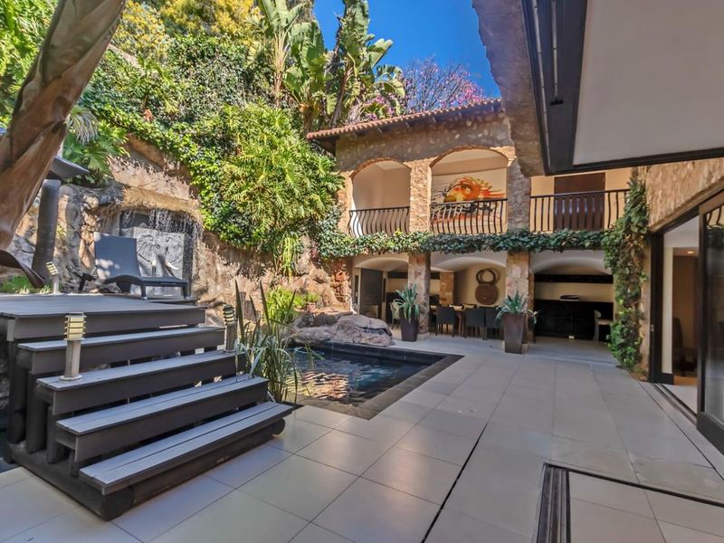 A beautifully landscaped outdoor pool area featuring stone arches, a built-in water feature, timber decking, and a spacious tiled patio for entertaining.