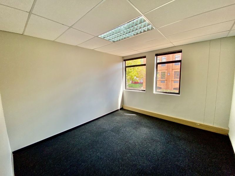 A clean, empty office space featuring dark carpeting, a suspended ceiling with recessed lighting, and large windows providing natural light.