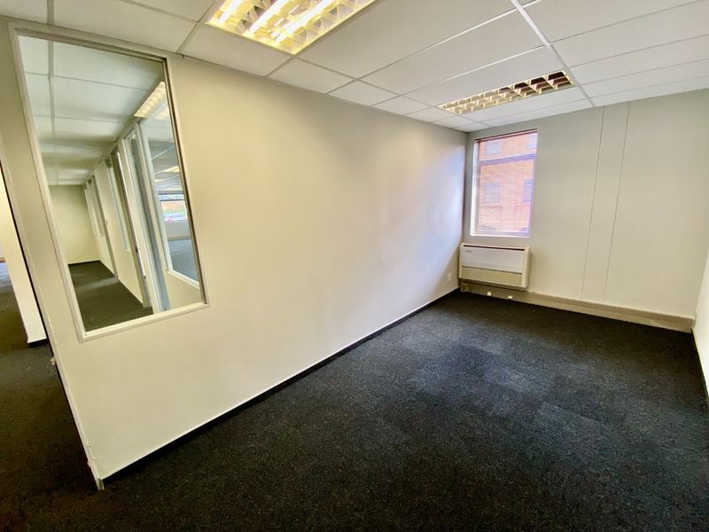 A vacant commercial office space featuring carpeted flooring, a suspended ceiling with recessed lighting, a glass partition wall, and a wall-mounted HVAC unit.