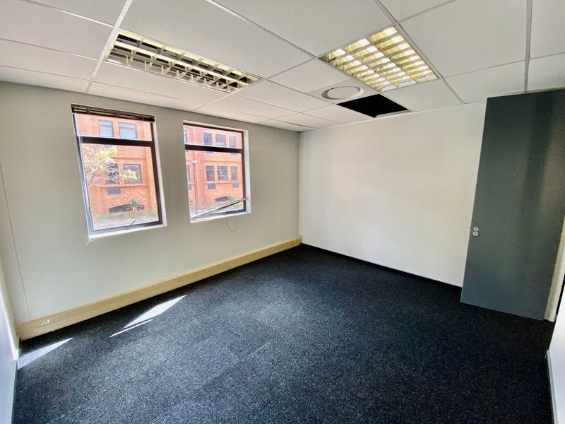 A vacant commercial office space featuring dark carpeting, suspended ceilings with recessed lighting, and large windows providing natural light.