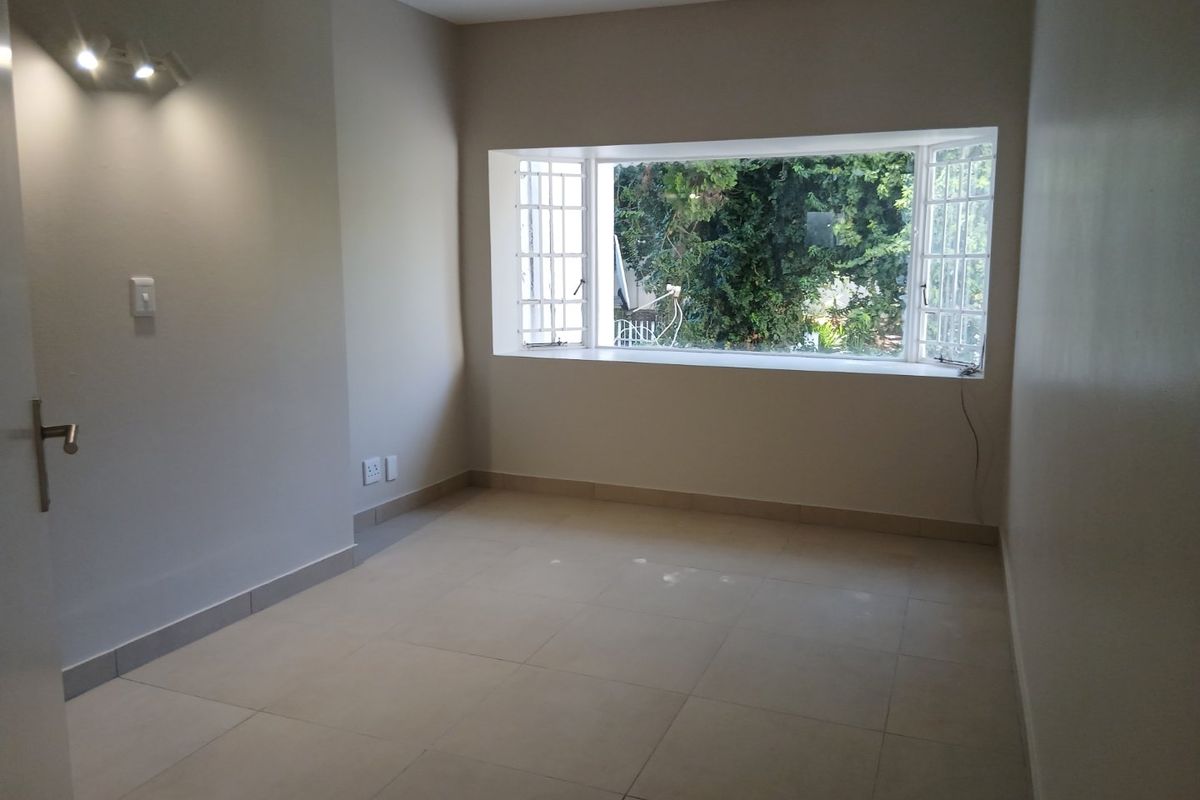 A bright, empty bedroom featuring large bay windows with security bars, neutral tiled flooring, and clean wall finishes.