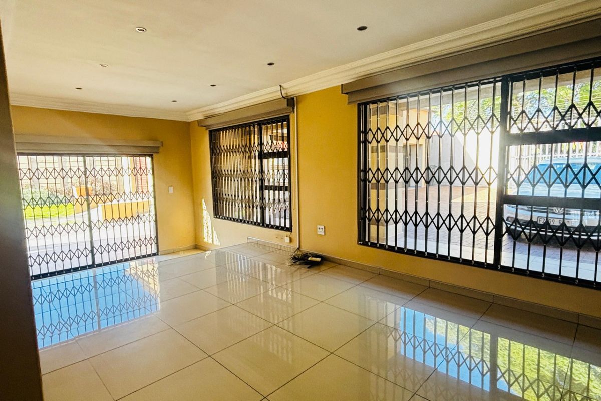 A spacious, empty living area featuring large windows with security gates, tiled flooring, and recessed ceiling lighting.