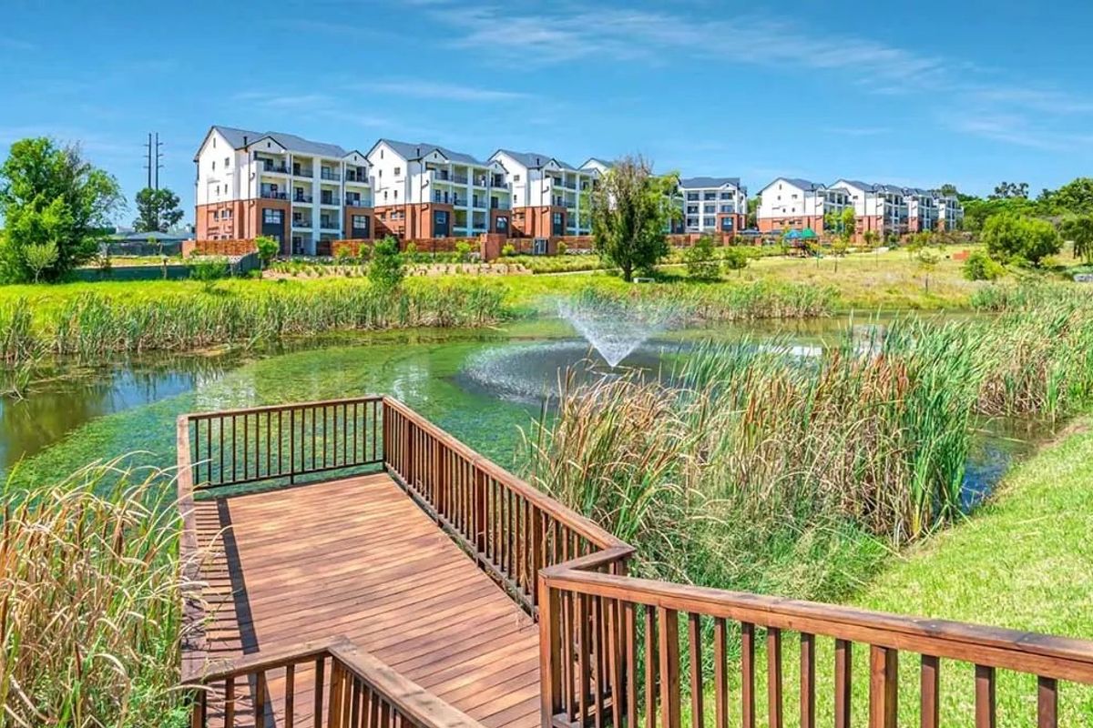 A scenic view of an apartment complex overlooking a landscaped pond with a wooden deck and water feature in the foreground.