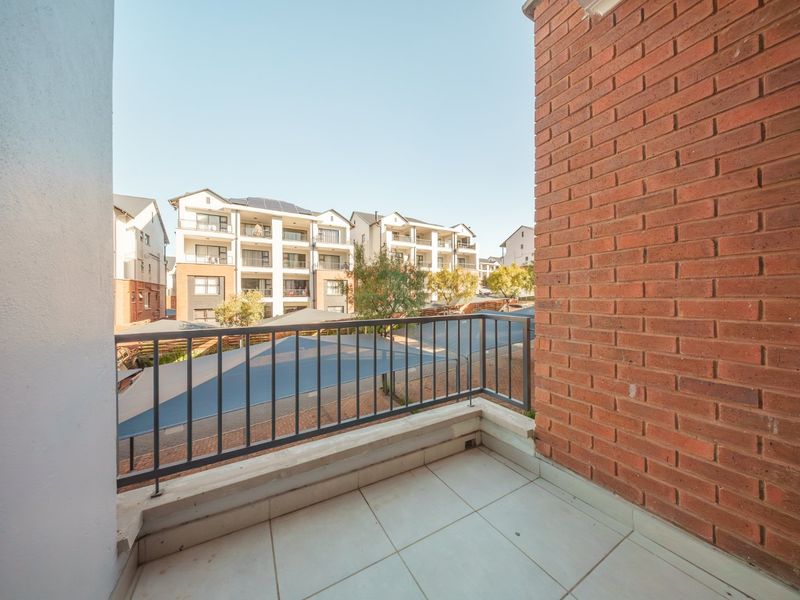 A clean, open balcony featuring tiled flooring, a metal balustrade, and an exposed brick wall, overlooking the apartment complex grounds.