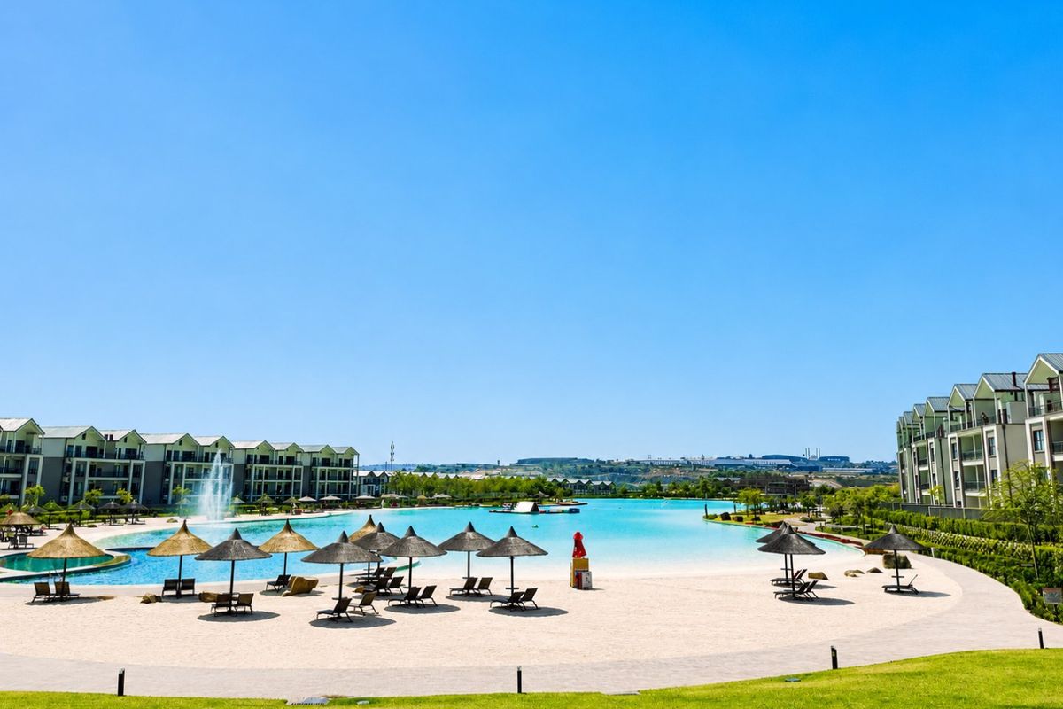 A large communal swimming pool area featuring a sandy beach entry, water fountain, and surrounding apartment buildings with landscaped grounds.