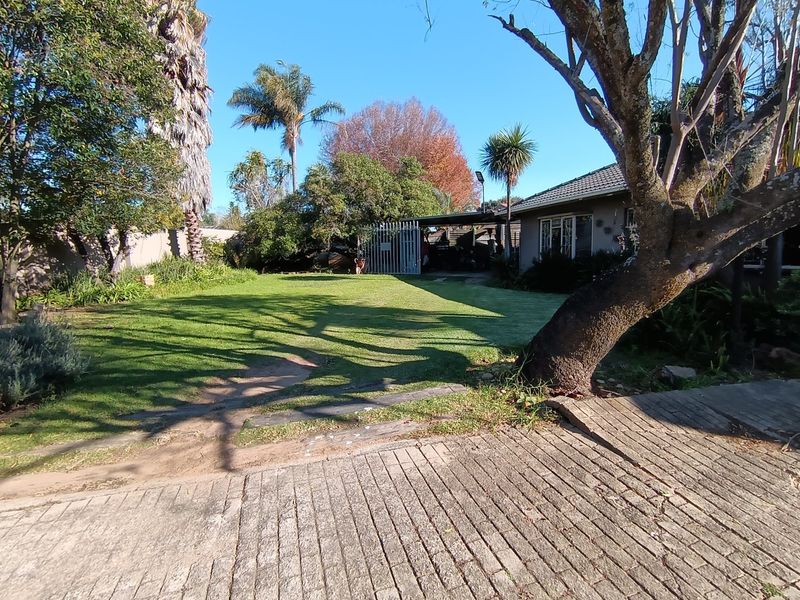Front view of the property featuring a paved driveway, established garden with mature trees, and a secure gated entrance.