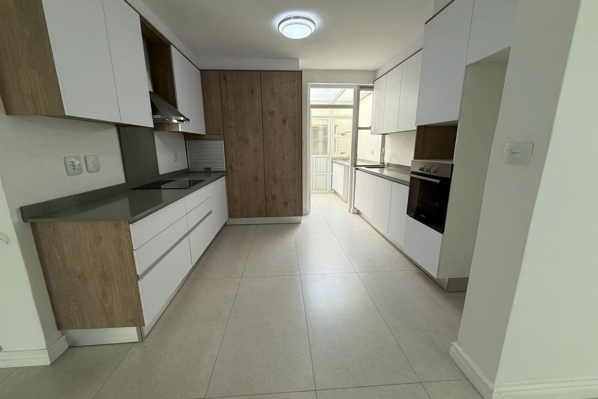 Modern galley-style kitchen featuring sleek white and wood-grain cabinetry, stone countertops, integrated oven, and hob, leading to a bright rear utility area.