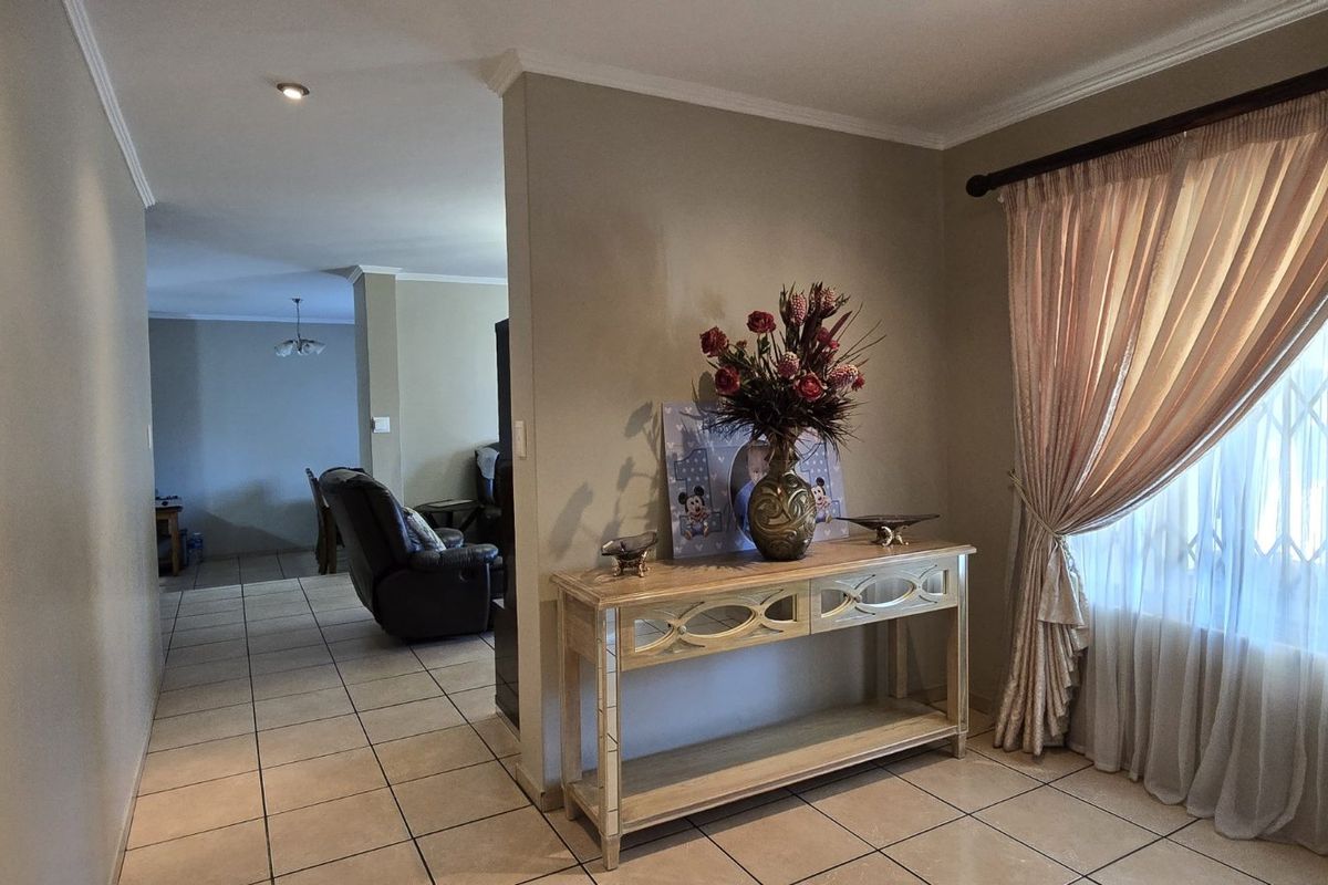 A bright living area featuring neutral tiled flooring, recessed lighting, and a large window with curtain treatments, leading into an adjacent open-plan space.