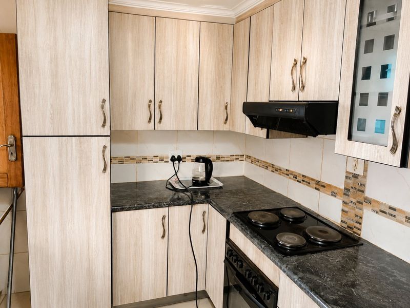 Compact kitchen featuring light wood-grain cabinetry, dark laminate countertops, built-in oven, electric hob, and a tiled backsplash.
