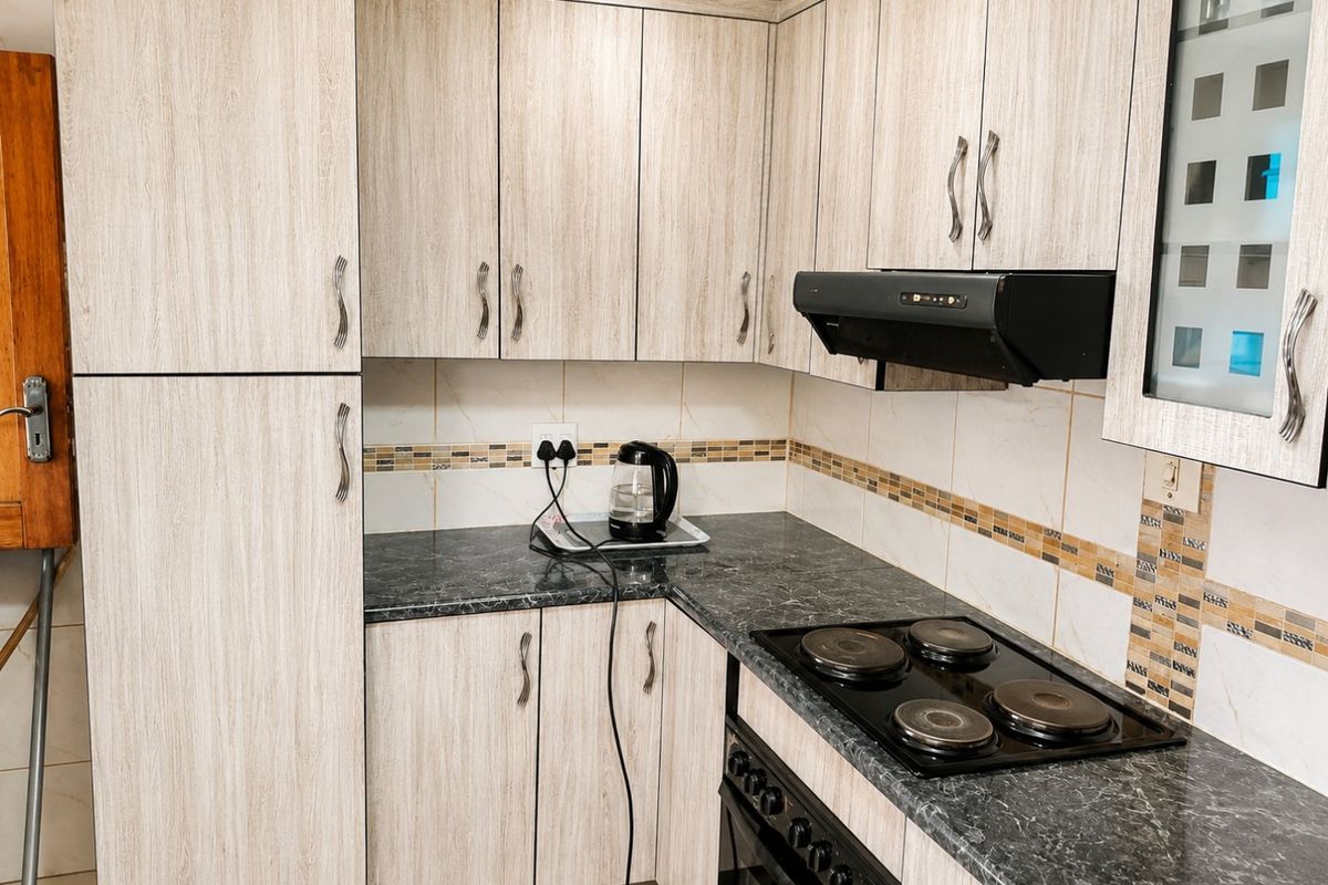 A compact kitchen featuring light wood-grain cabinetry, dark laminate countertops, a built-in oven, electric hob, and a tiled backsplash.